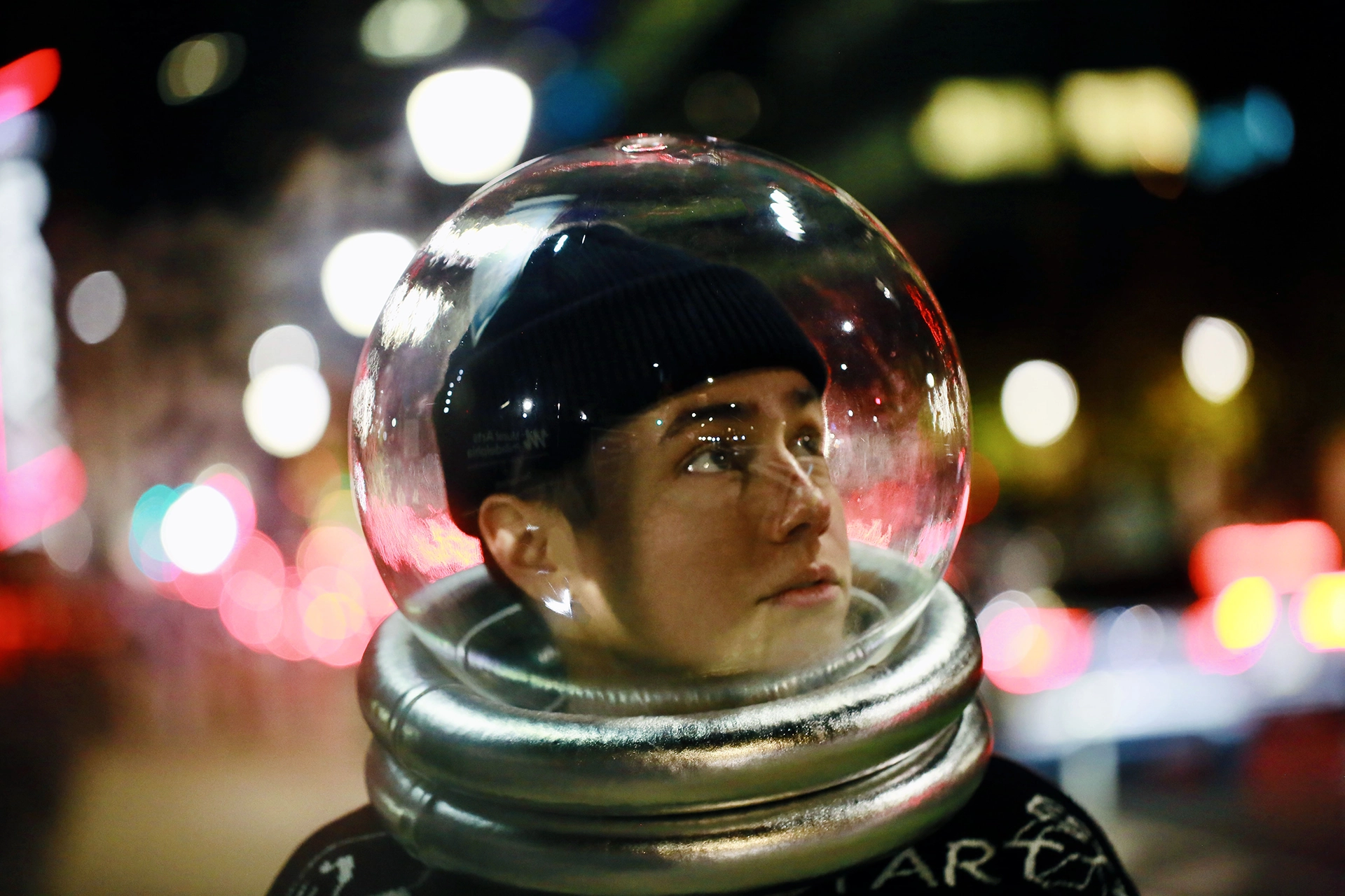 Person wearing a transparent space helmet with a black beanie underneath, looking up against a blurred city night background with colorful lights.