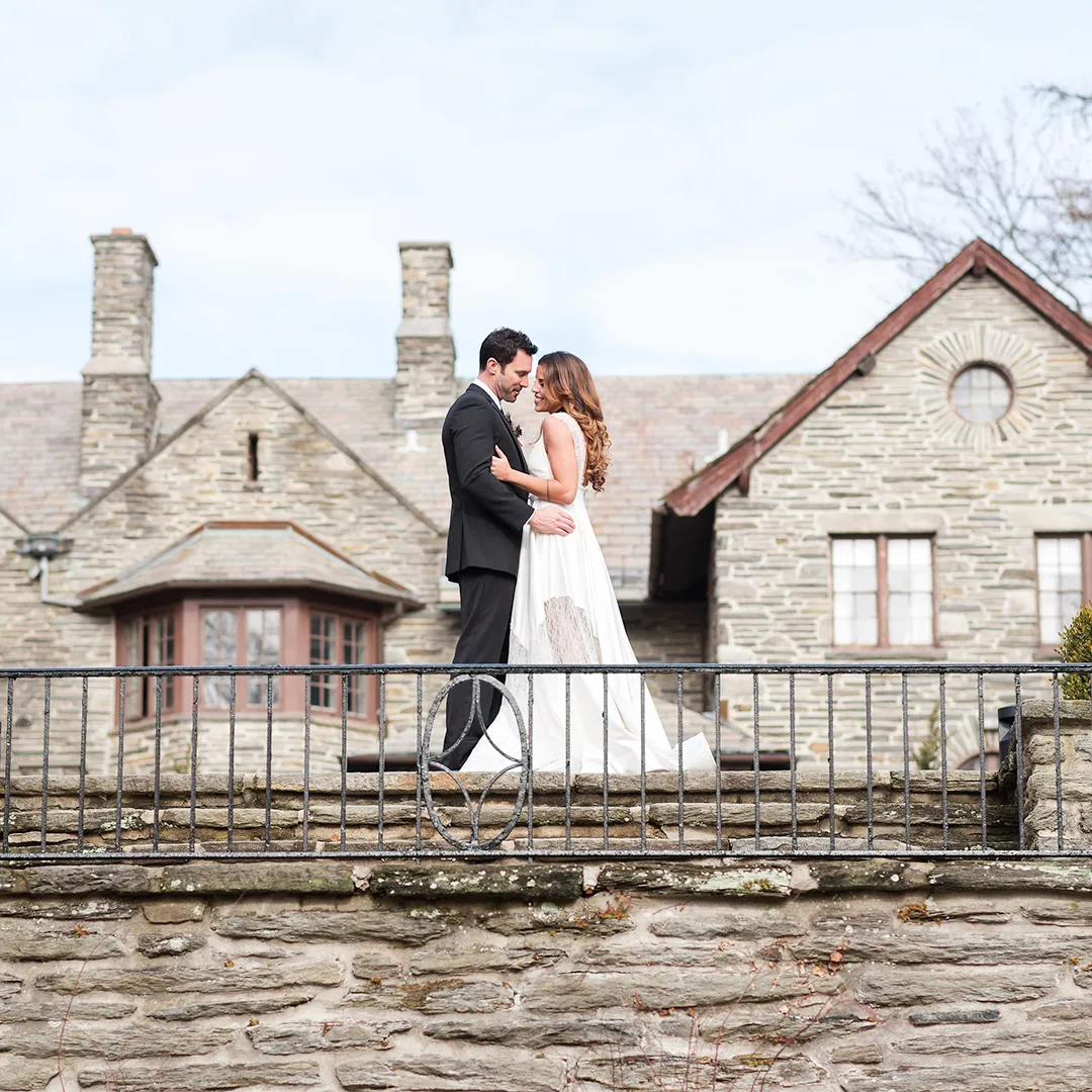 Bride and groom embracing on a stone terrace in front of a large stone house.