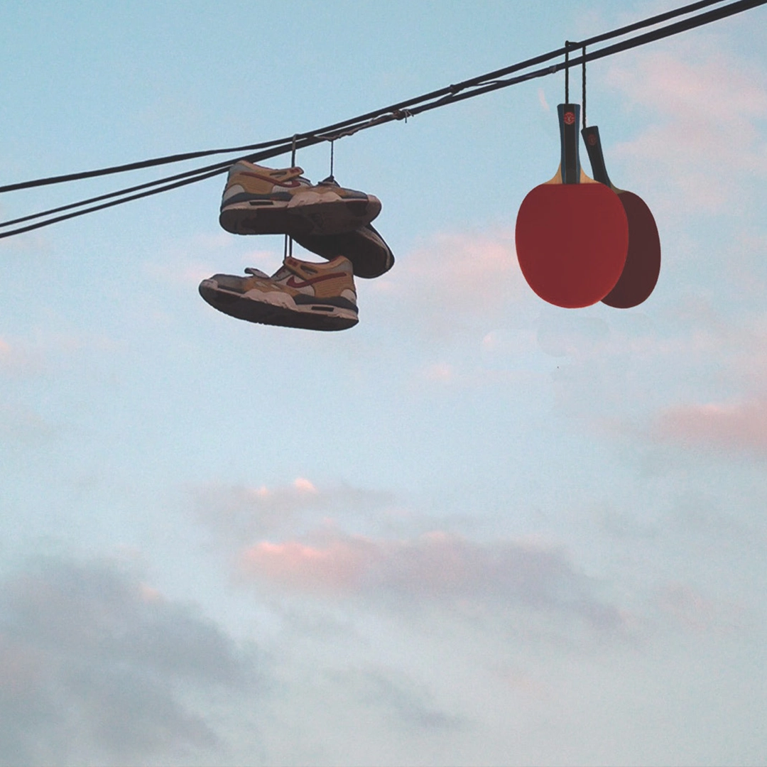 Pair of sneakers and two red ping pong paddles hanging on a wire against a sky with scattered clouds.