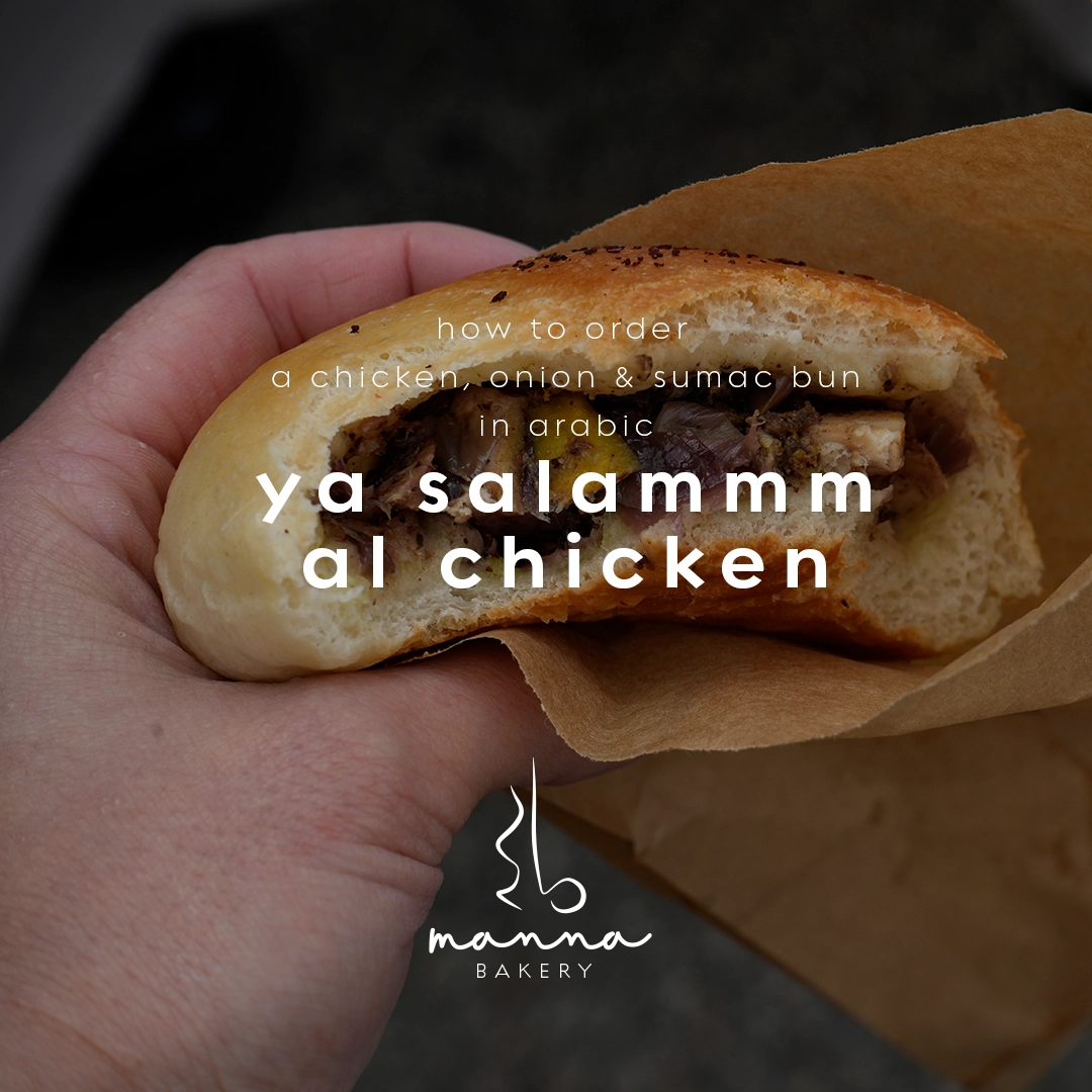 Hand holding a half-eaten chicken, onion, and sumac bun wrapped in brown paper, with text showing Arabic ordering phrase for the bun.