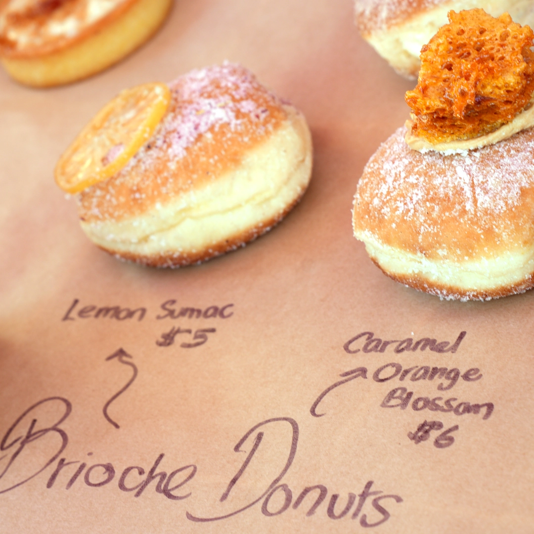 Two brioche donuts on brown paper, one topped with candied lemon slice labeled Lemon Sumac $5, the other with caramel orange blossom priced $6.