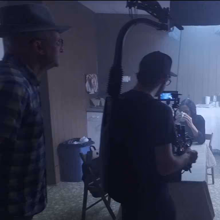 Film crew member operating a camera rig while a woman sits at a table in a dimly lit room.