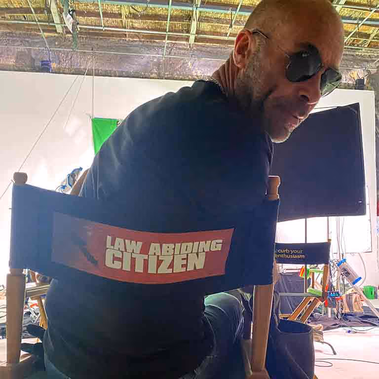 Man wearing sunglasses sitting on a director's chair labeled 'Law Abiding Citizen' on set with film equipment and another chair labeled 'Curb Your Enthusiasm' in the background.